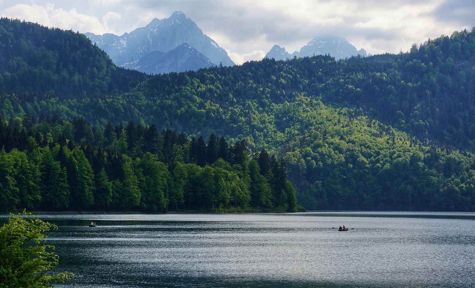 A serene lake surrounded by lush green forests and mountains in the background under a cloudy sky.